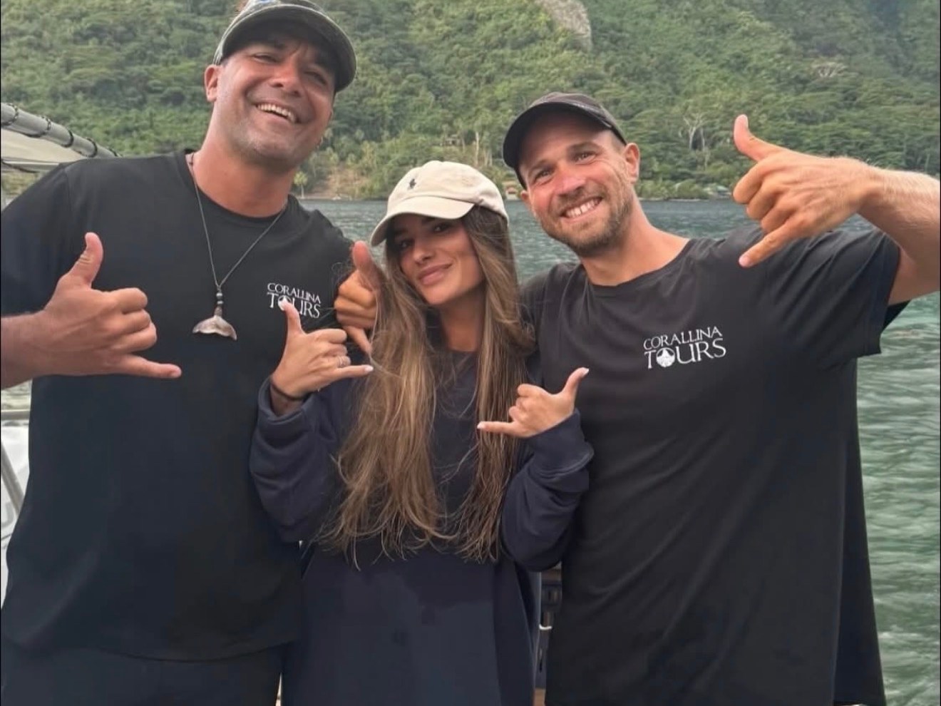 Three people posing on a boat with mountains in the background, making 'shaka' hand signs.