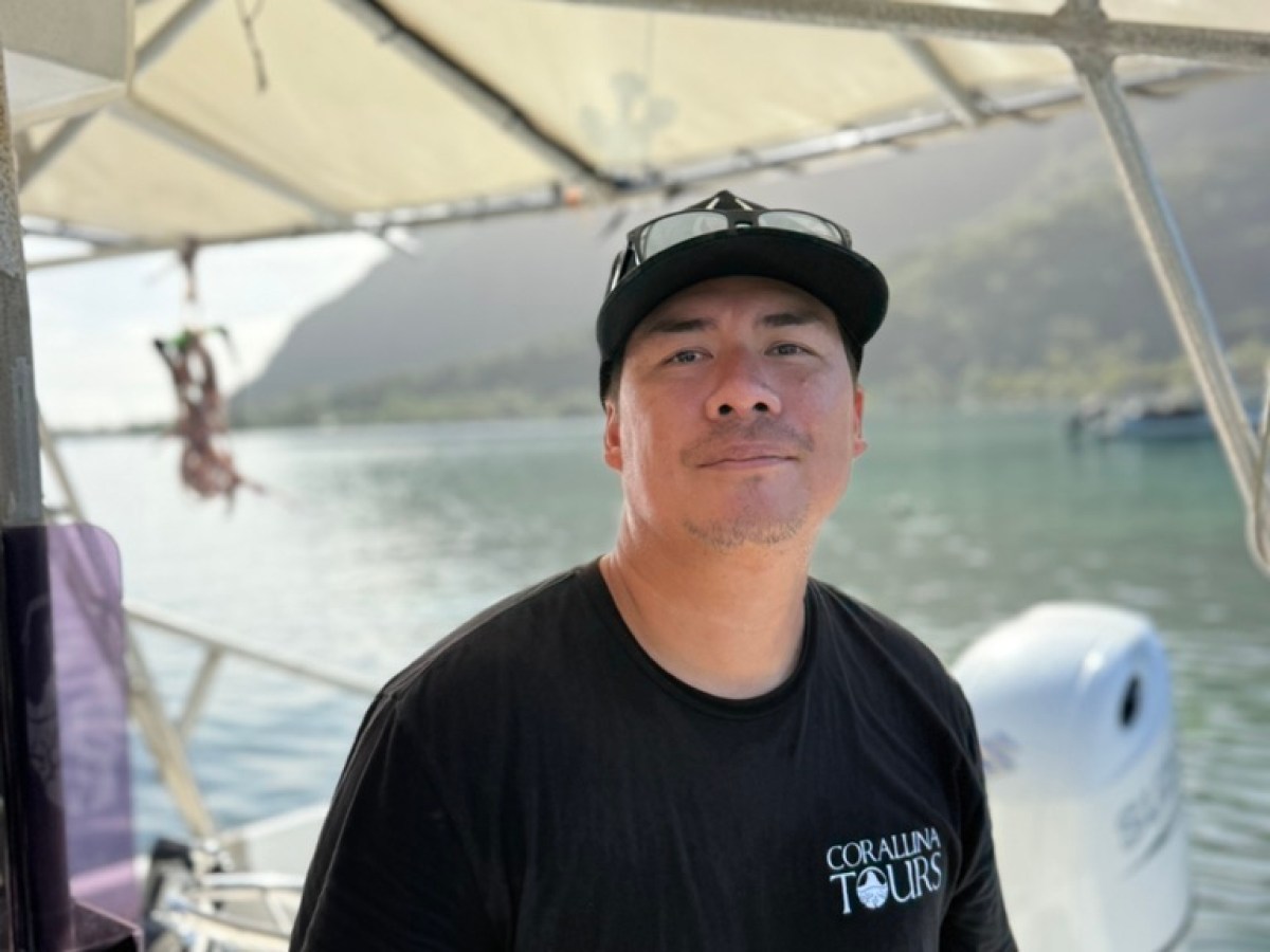 Man on a boat wearing a black shirt and cap, with water and mountains in the background.